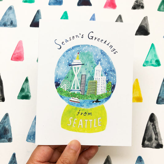 Watercolor greeting card featuring a Seattle snow globe with iconic landmarks, surrounded by colorful triangles. Hand holding the card, showcasing "Season's Greetings from Seattle" text and vibrant cityscape inside the globe.