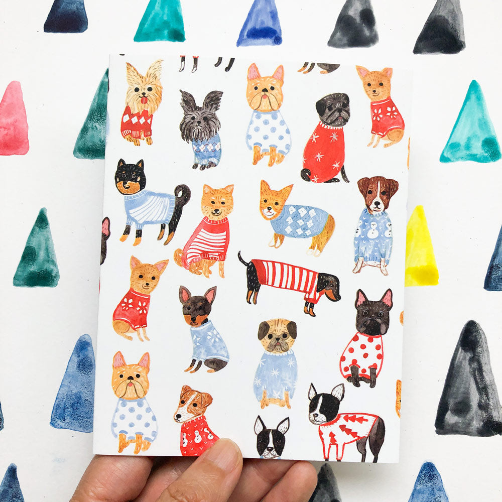 Colorful holiday greeting card featuring various small dog breeds wearing festive sweaters and patterns, held by a hand against a background of painted triangular shapes