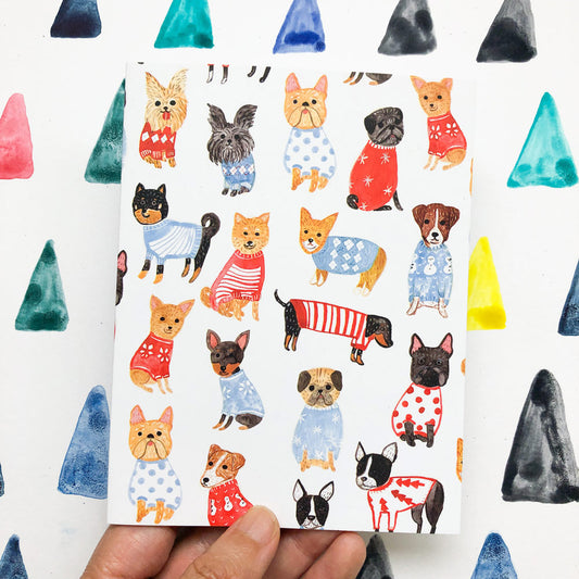 Colorful holiday greeting card featuring various small dog breeds wearing festive sweaters and patterns, held by a hand against a background of painted triangular shapes