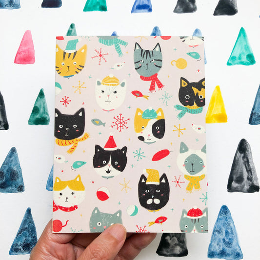 Festive holiday greeting card featuring colorful cartoon cat faces with winter accessories, surrounded by snowflakes and stars, held against a background of watercolor triangles