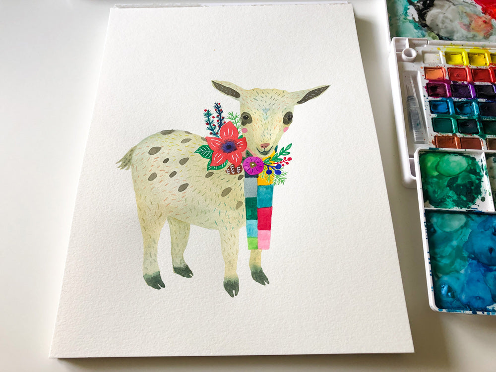 Watercolor painting of a baby goat with colorful floral decorations, standing on a white greeting card, with paint palette visible in corner