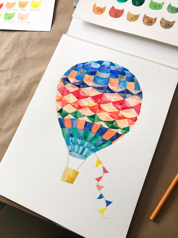 Colorful watercolor hot air balloon greeting card with geometric pattern and festive flags, displayed on kraft paper alongside cat-themed color swatches and an orange pencil