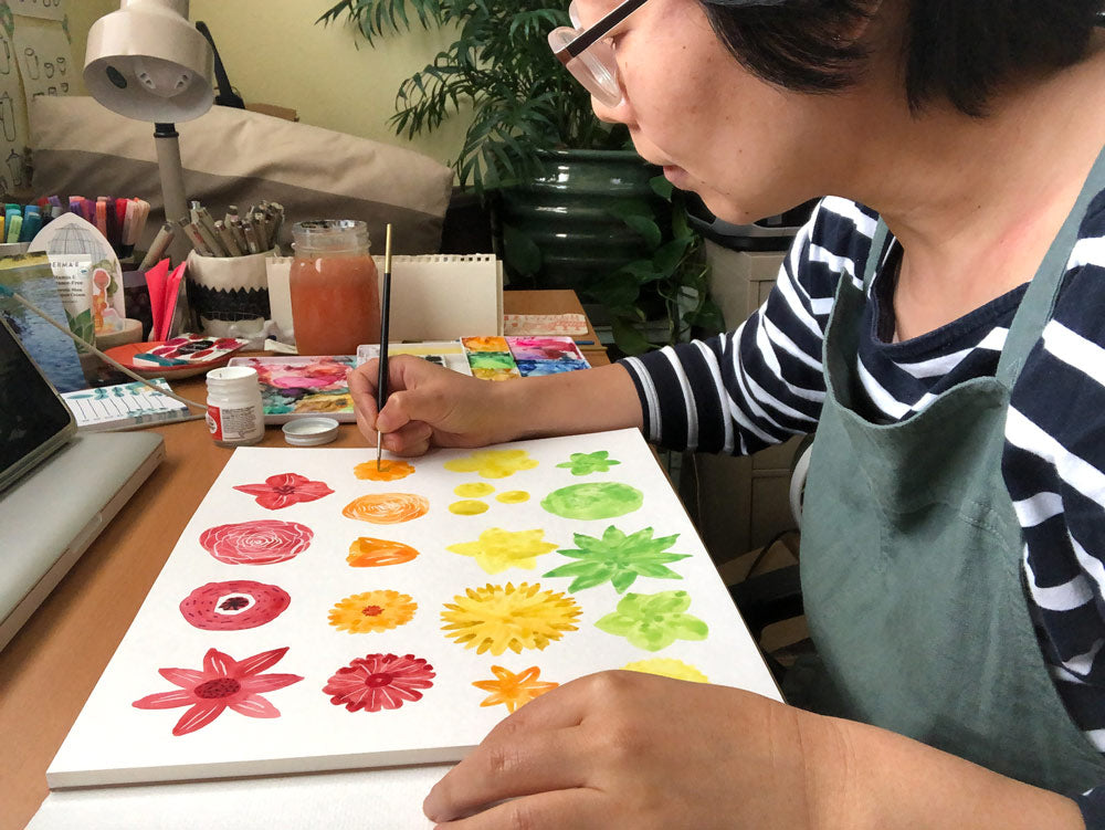 Artist painting colorful rainbow flower chart in watercolor on white paper, surrounded by art supplies on a desk with lamp and plants in background