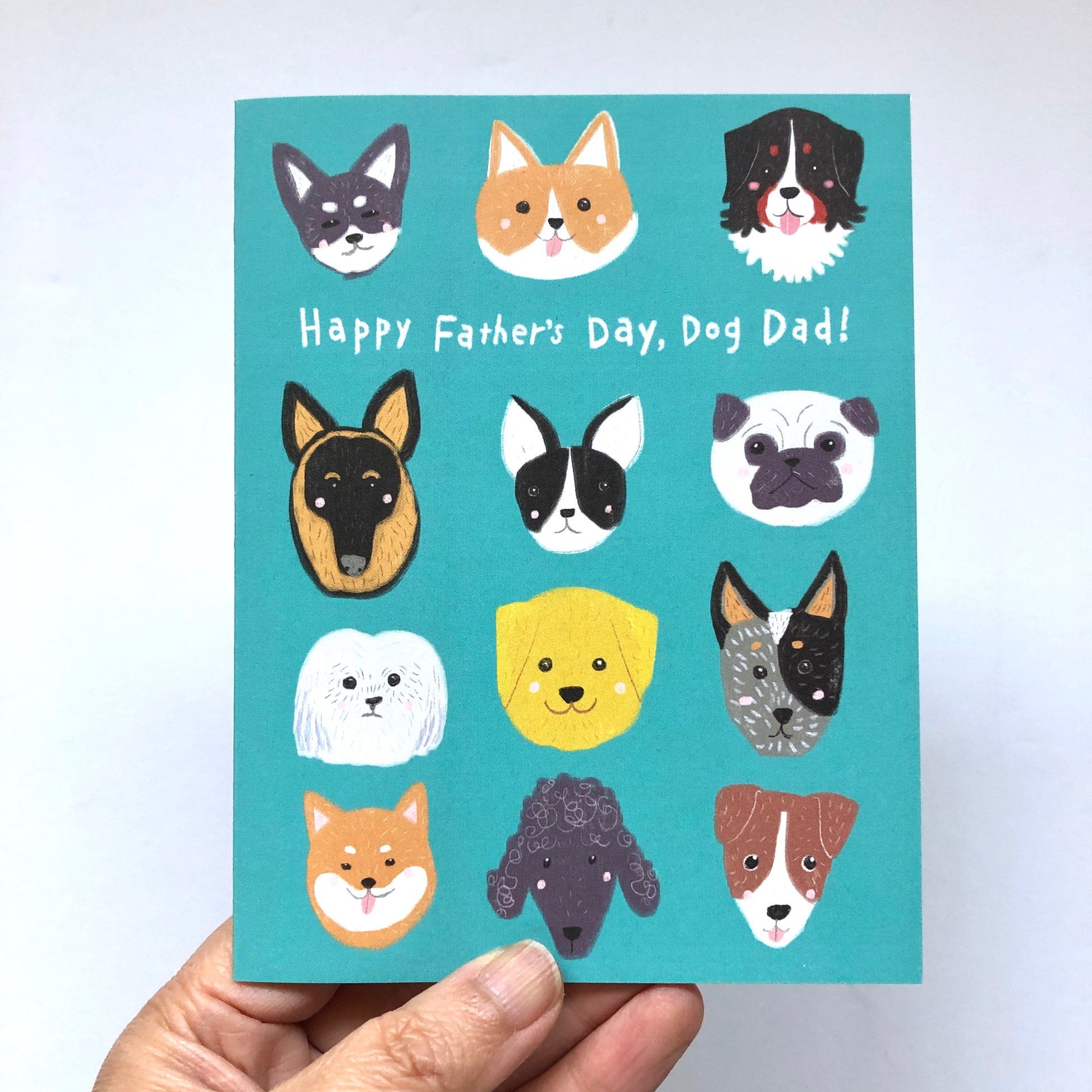 Teal Father's Day card featuring diverse dog breed faces with text "Happy Father's Day, Dog Dad!" Includes various breeds like Husky, Corgi, Bernese Mountain Dog, German Shepherd, Boston Terrier, Pug, and more. Hand holding the card visible.