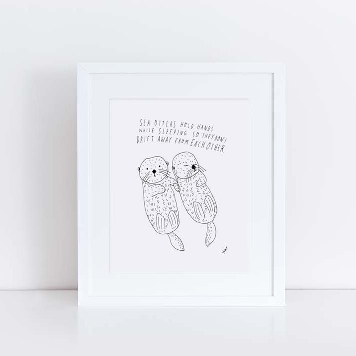 Framed pen and ink illustration of two sea otters holding hands while floating, with handwritten text above describing their sleeping habit to stay together