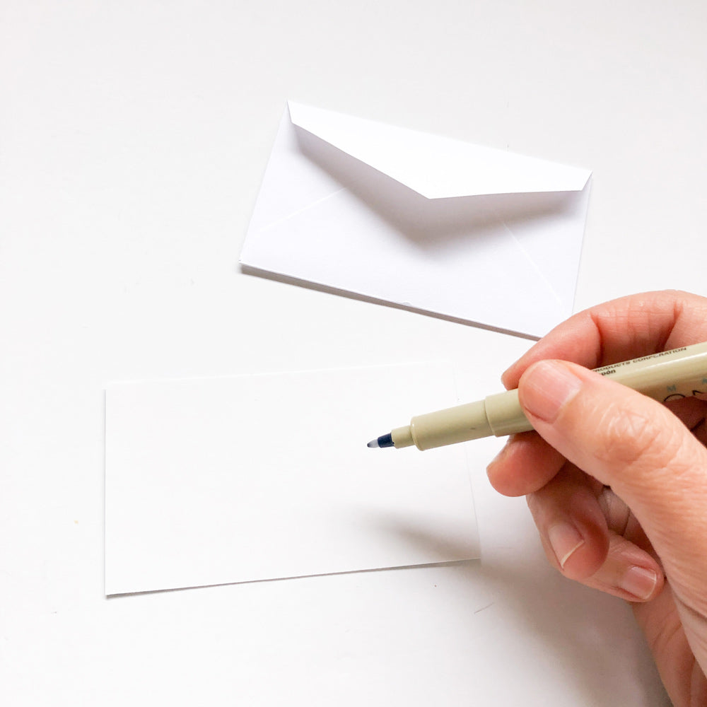 Hand holding pen over blank white card with matching envelope, showcasing tiny note set for personalized messages and gift enclosures