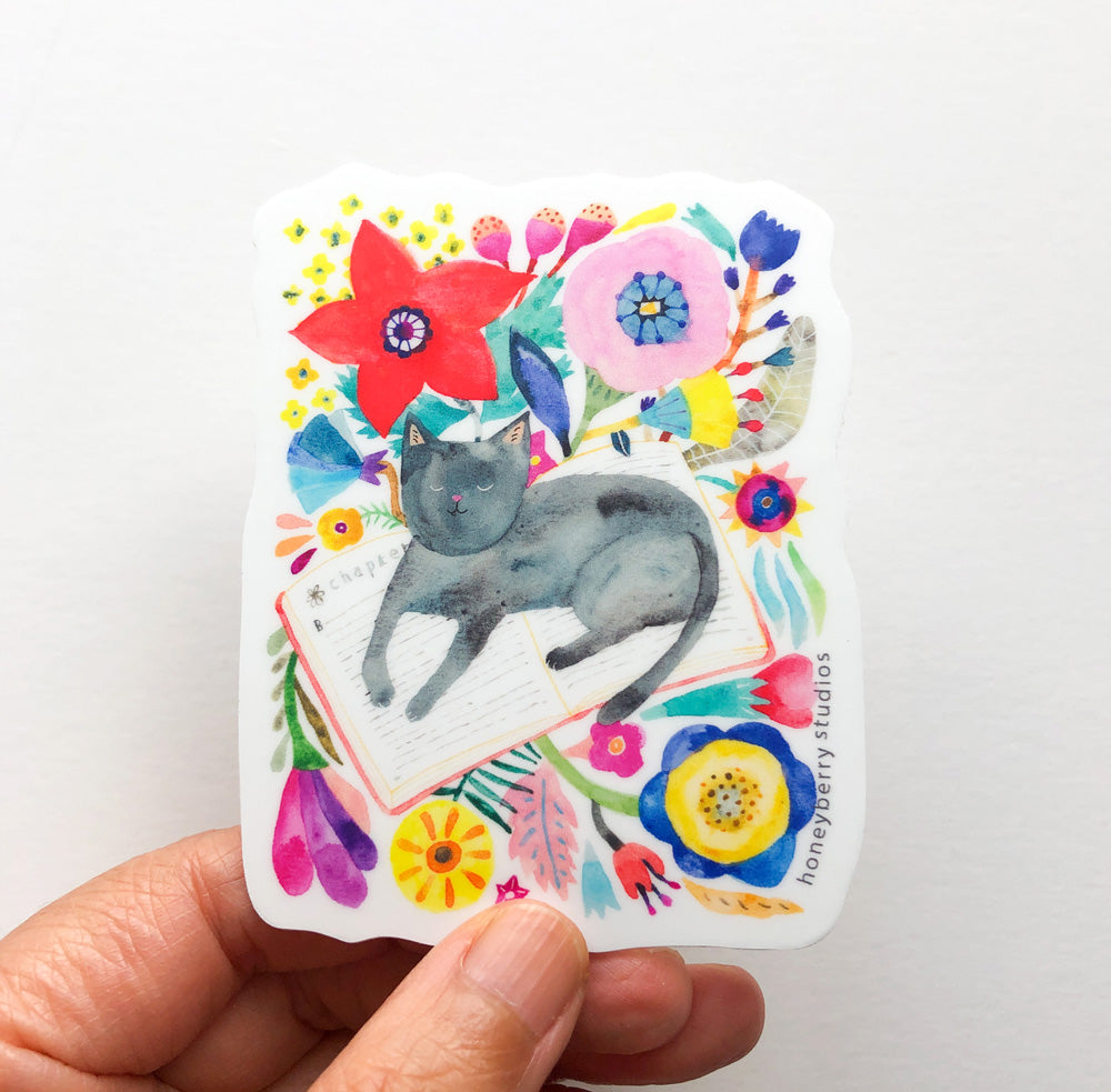 Colorful watercolor sticker featuring a gray cat napping on an open book, surrounded by vibrant flowers and foliage in red, pink, blue, and yellow. Hand holding the sticker to show size and design.