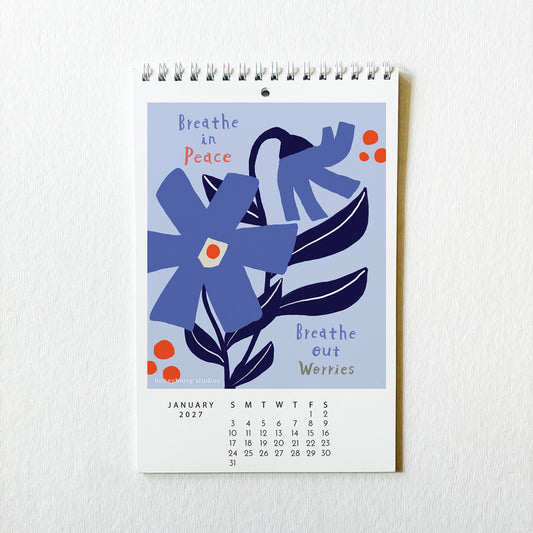 A spiral-bound January 2027 desk calendar hanging on a textured white wall. The cover features a minimalist illustration of blue flowers and leaves with the text 'Breathe in Peace' and 'Breathe out Worries' on a light blue background.