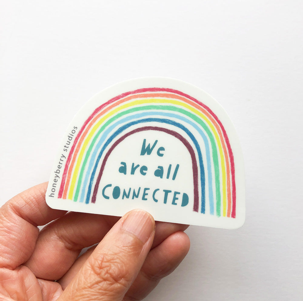 Colorful rainbow-shaped vinyl sticker with hand-lettered text "We are all connected" held between fingers, showcasing size and design of inspirational Shopify product