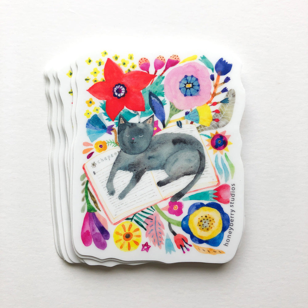 Watercolor sticker featuring a gray cat napping on an open book surrounded by colorful flowers and foliage, perfect for cat lovers and book enthusiasts