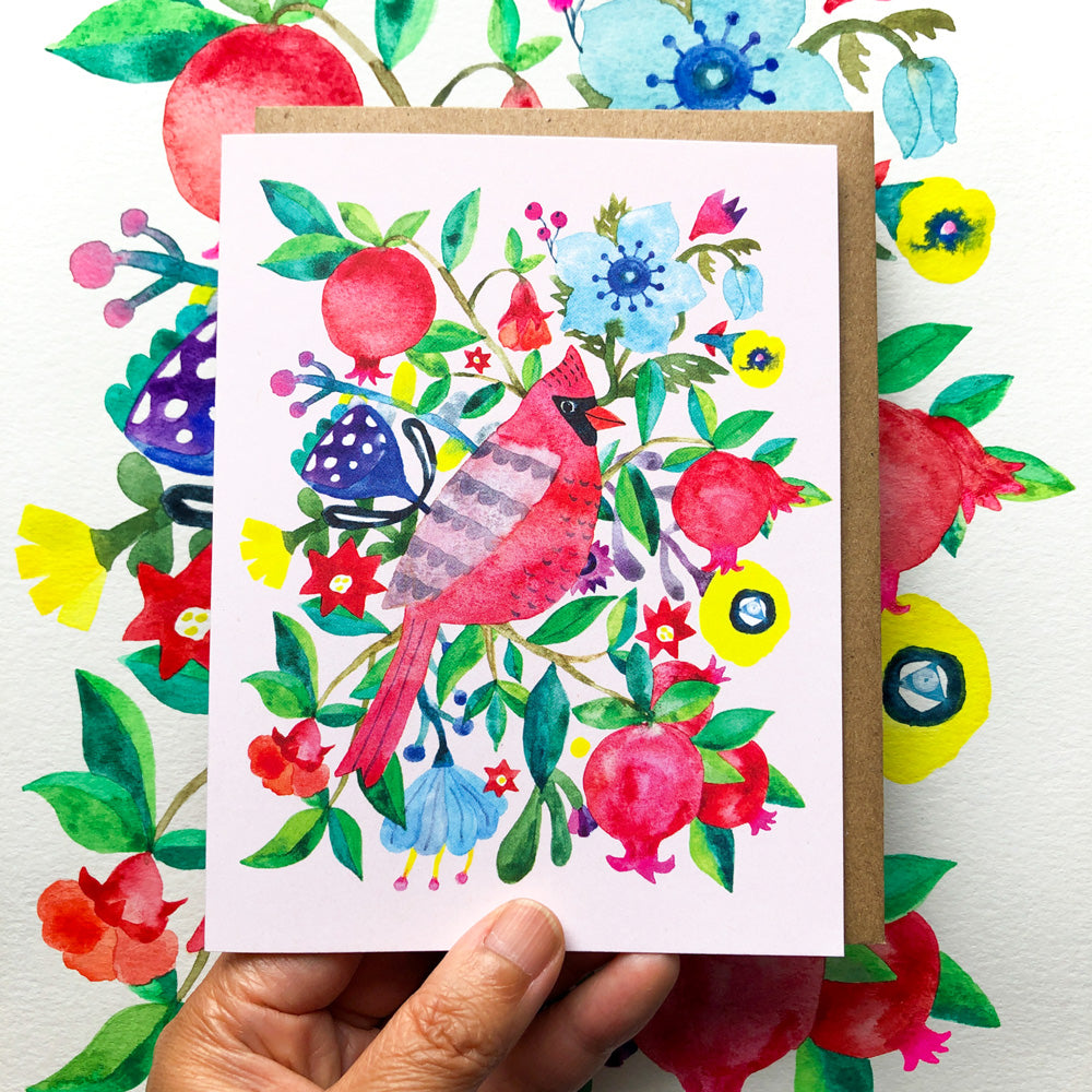 Colorful watercolor greeting card featuring a vibrant red cardinal bird surrounded by pomegranates, flowers, and festive holiday elements on a white background, held by a hand against a floral patterned surface