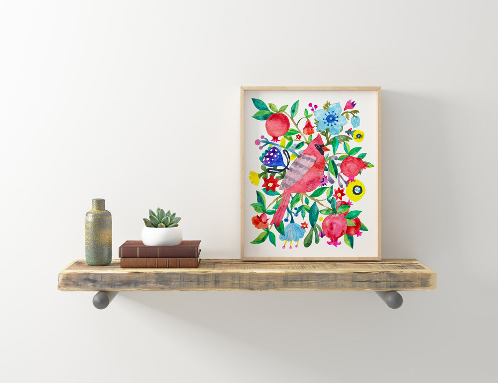 Colorful giclee art print featuring a vibrant cardinal and pomegranate surrounded by flowers and foliage, displayed on a rustic wooden shelf with decorative items, showcasing the 8x10 inch watercolor paper artwork in a light wooden frame