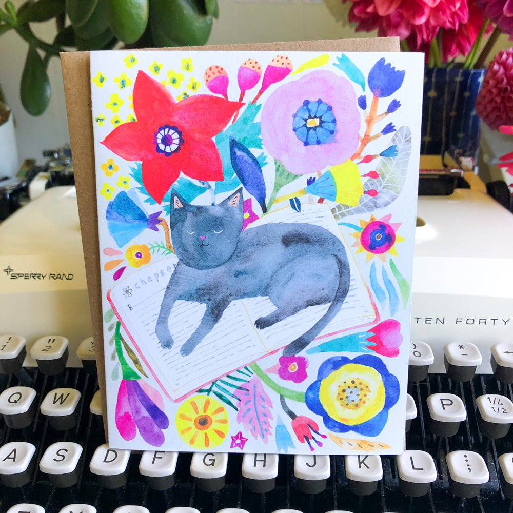 Colorful watercolor greeting card featuring a gray cat napping among vibrant flowers and leaves on white background, displayed on a vintage typewriter