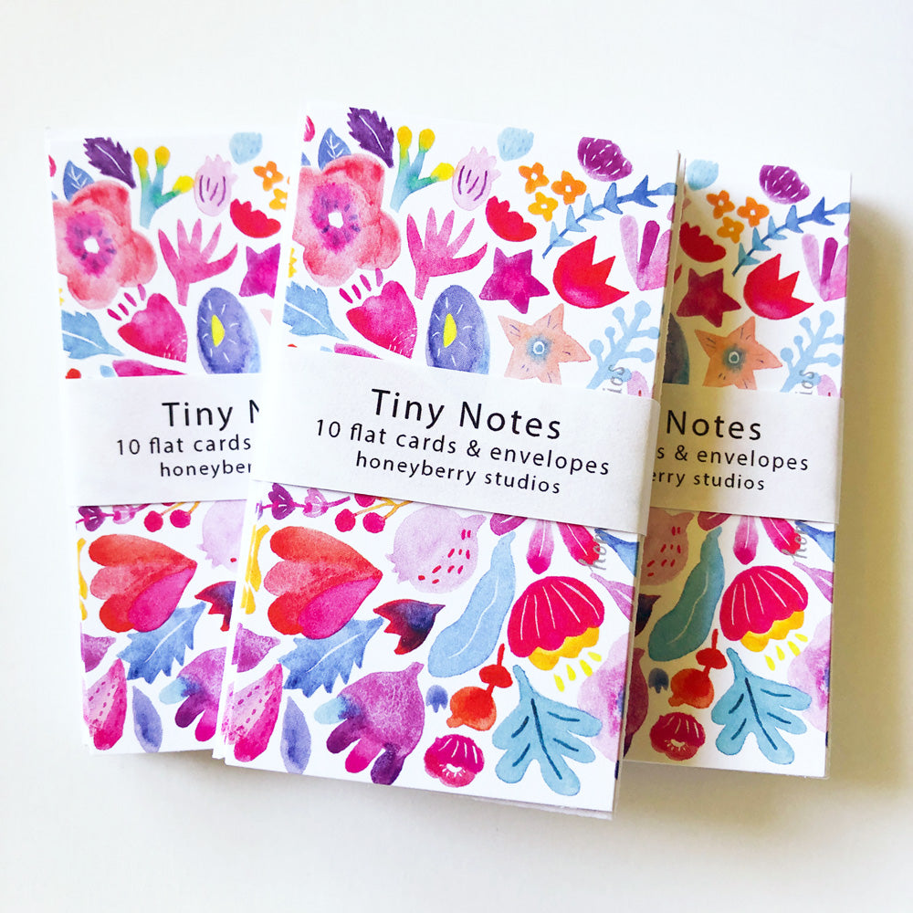 Colorful watercolor floral tiny notes sets featuring vibrant red, pink, and purple flowers with blue leaves. Each pack contains 10 flat cards and envelopes, wrapped with a band labeled "Tiny Notes" from Honeyberry Studios.