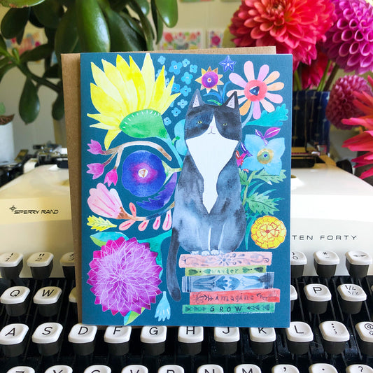 Colorful greeting card featuring a watercolor illustration of a tuxedo cat sitting on books, surrounded by vibrant flowers and plants, displayed on a vintage typewriter with a pink dahlia in the background