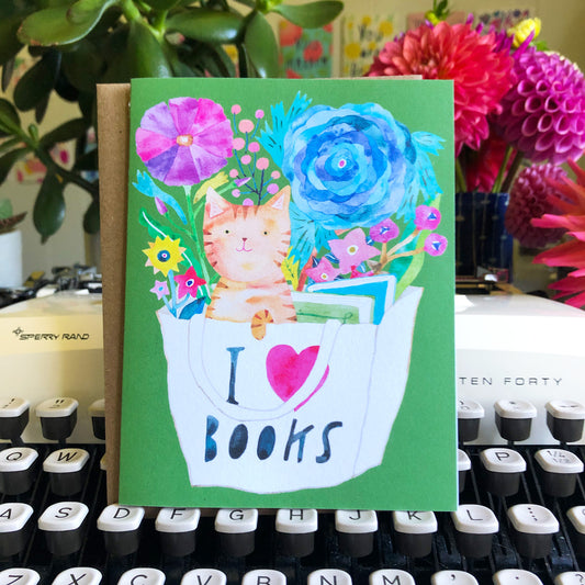 Colorful greeting card featuring a watercolor illustration of an orange tabby cat in an 'I love books' bag, surrounded by vibrant flowers on a green background, displayed on a typewriter