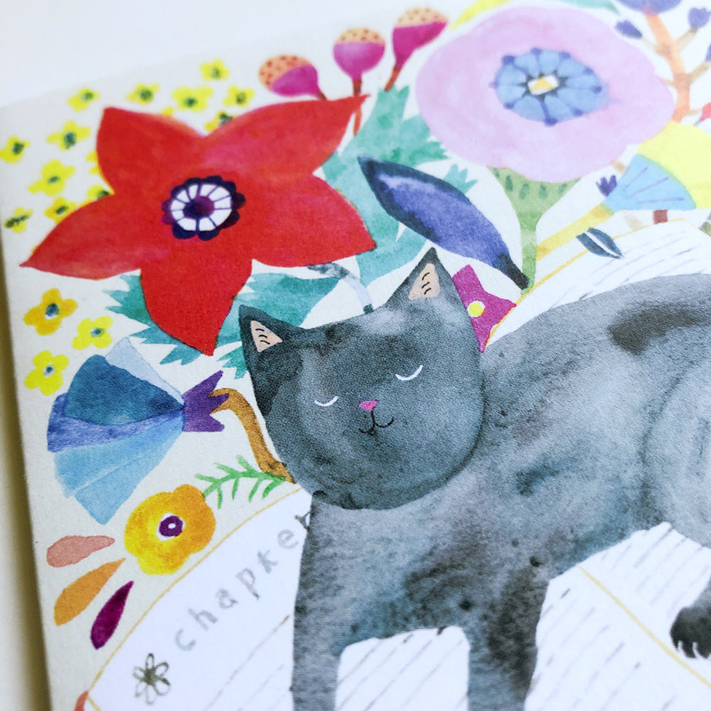 Watercolor greeting card featuring a sleeping gray cat on books surrounded by vibrant flowers, including a large red bloom and pastel pink and yellow blossoms, creating a whimsical and colorful design