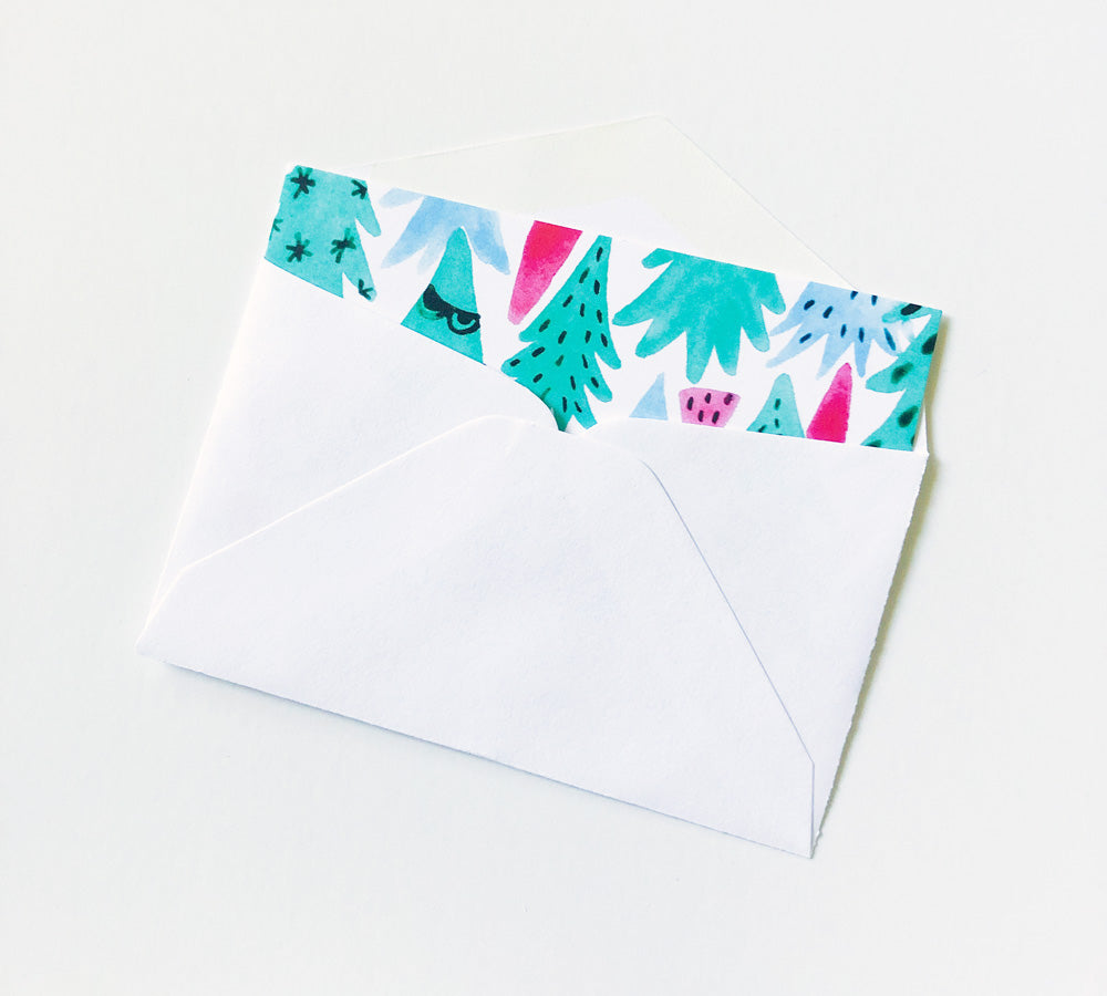 White envelope with colorful Christmas tree design peeking out from the top, featuring teal, pink, and blue watercolor illustrations of festive trees and shapes