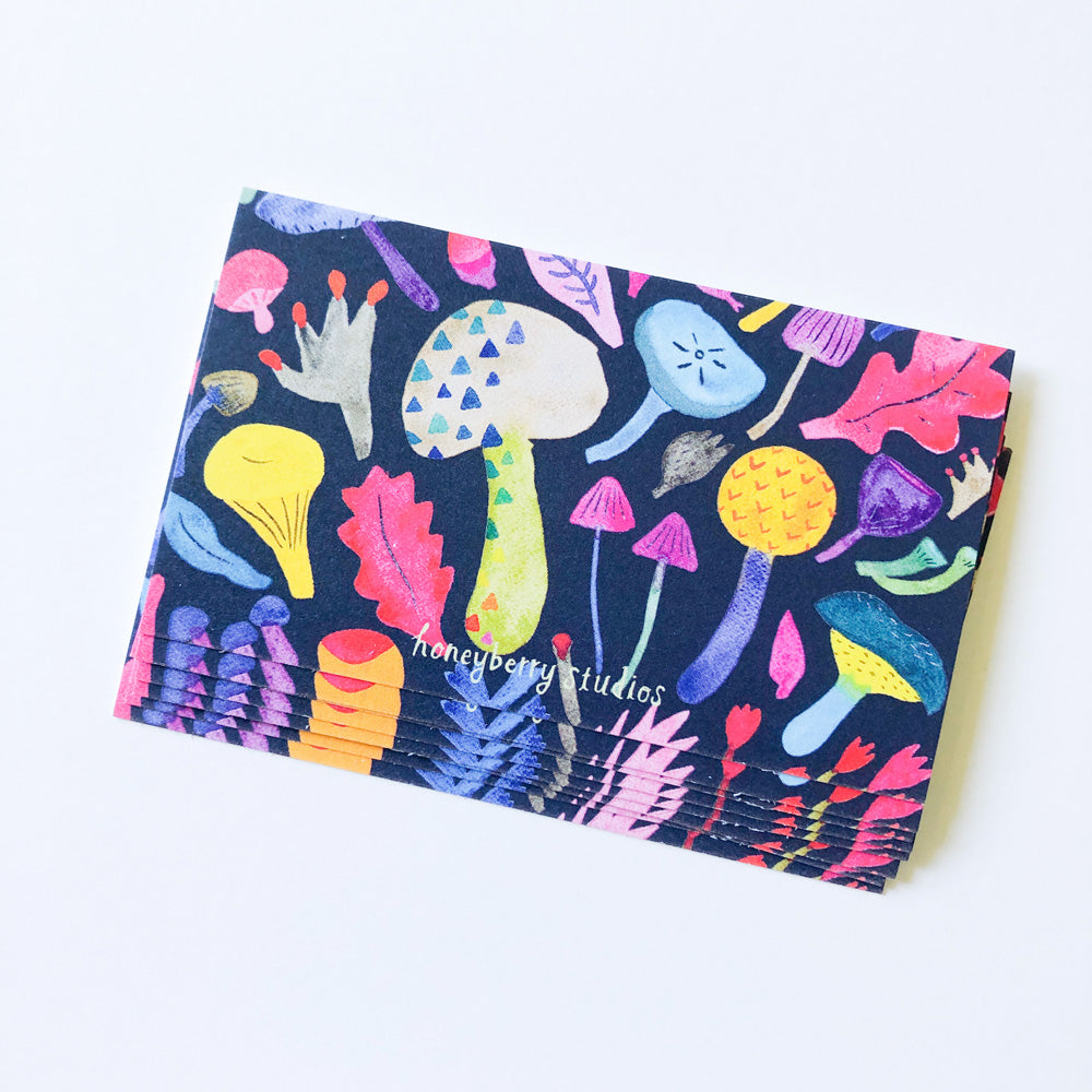 Colorful watercolor mushroom illustration on dark blue background, featuring various whimsical shapes and sizes of fungi in vibrant hues, displayed on a small rectangular notecard for gift enclosures or tiny greetings.