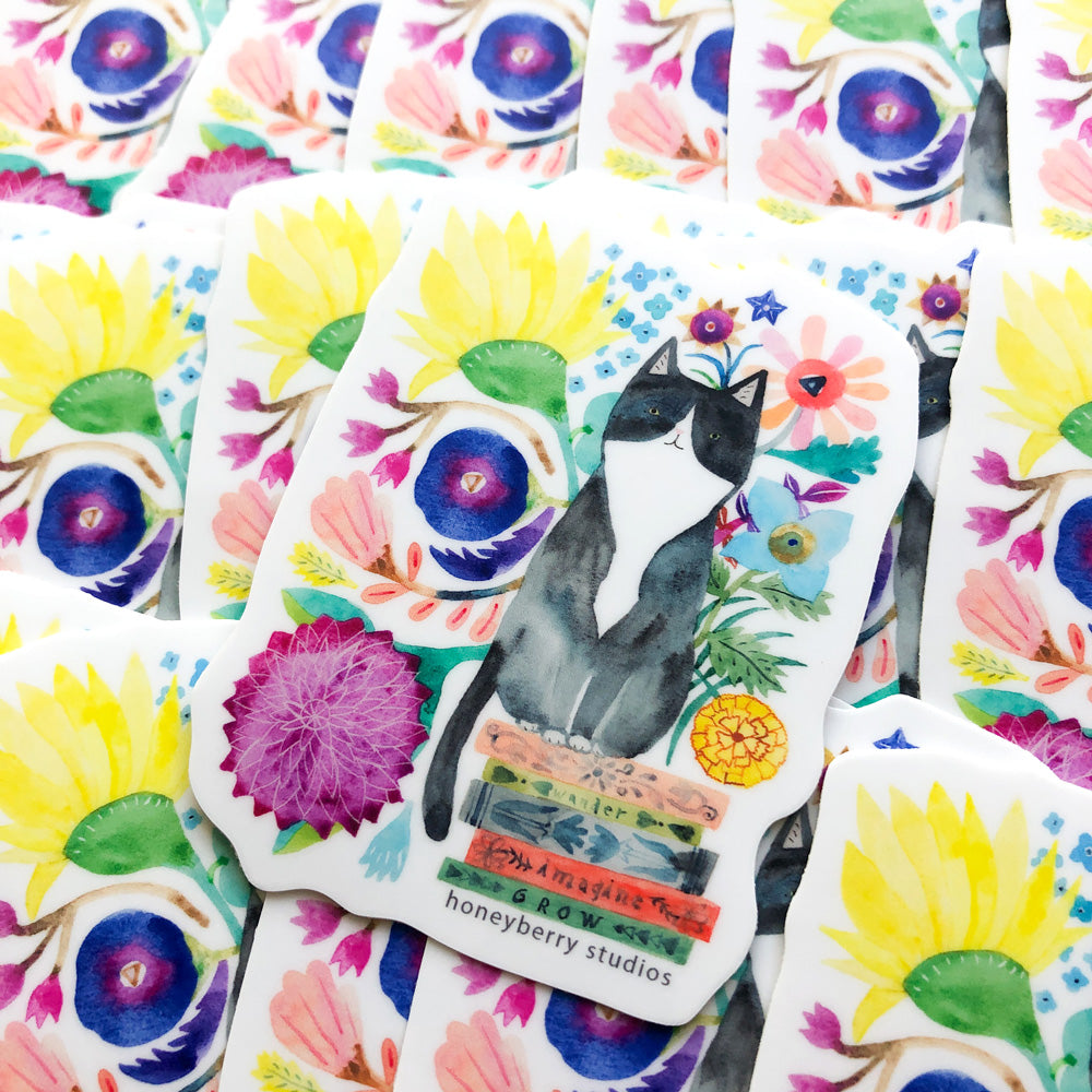 Watercolor vinyl sticker featuring a tuxedo cat sitting on books, surrounded by colorful flowers and a pineapple, placed on a floral background with yellow sunflowers and purple blooms