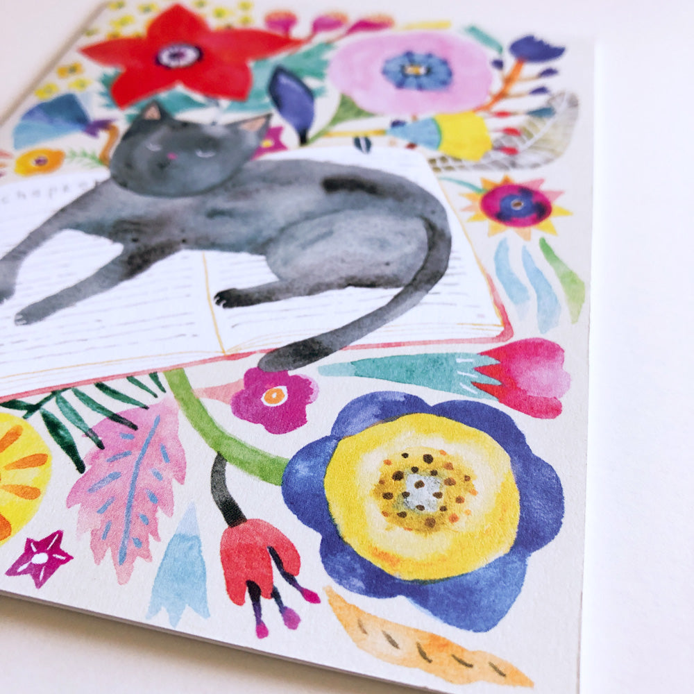 Watercolor greeting card featuring a gray cat napping on open books, surrounded by vibrant floral designs including red, pink, blue, and yellow flowers, leaves, and abstract shapes on a white background