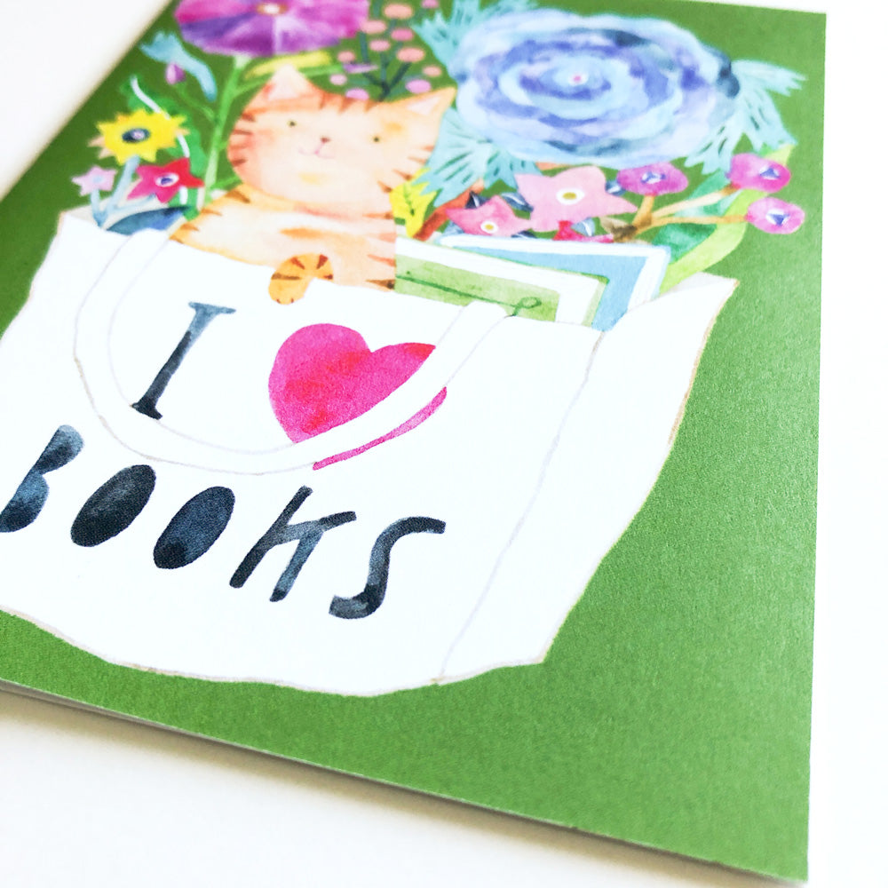 Colorful greeting card featuring a watercolor illustration of an orange tabby cat peeking out of a white book bag with 'I love books' text, surrounded by vibrant flowers on a green background