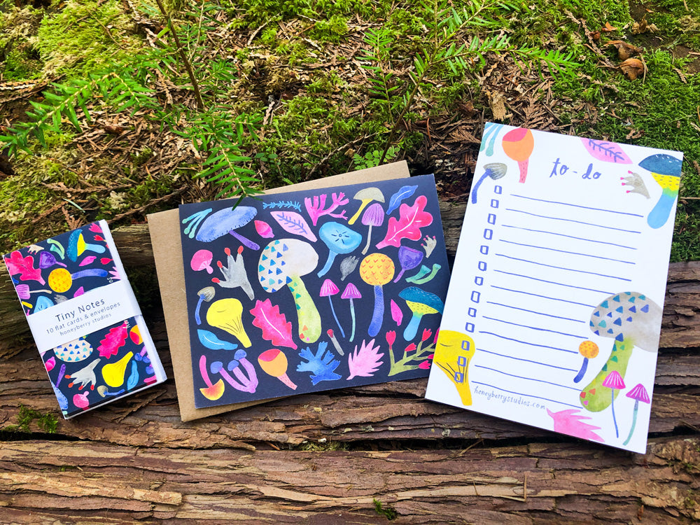 Colorful mushroom-themed stationery set displayed on moss-covered wood, featuring a to-do list notepad, greeting card, and tiny notes with vibrant watercolor mushroom illustrations on dark backgrounds