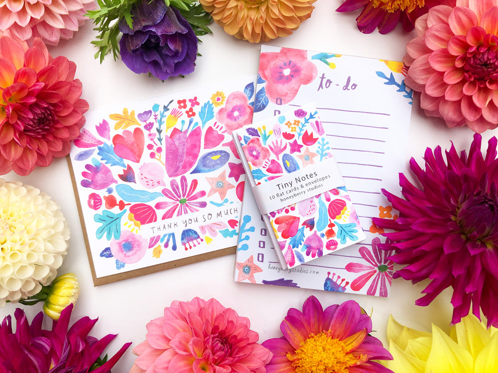 Colorful floral to-do list notepad surrounded by vibrant real flowers, including dahlias and anemones, on a white background. The notepad features a cheerful pink and red flower design matching the surrounding blooms.