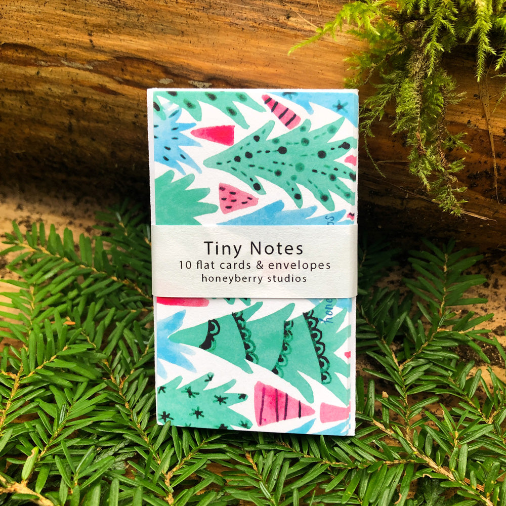 Colorful Christmas forest tiny notes set on wooden surface surrounded by green ferns, featuring watercolor illustrations of stylized trees in festive red and green tones