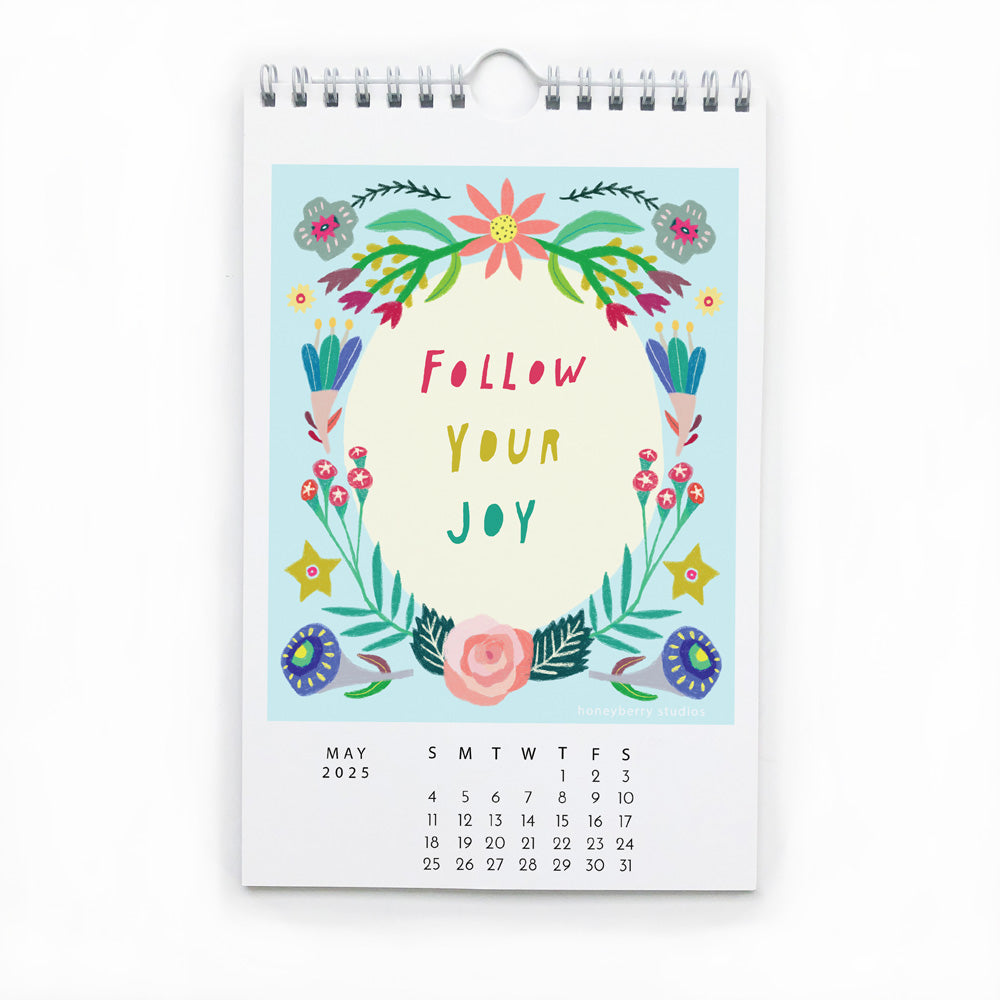 Colorful wall calendar page for May 2025 featuring floral illustration and hand-lettered "Follow Your Joy" message, with spiral binding and white wire hanger