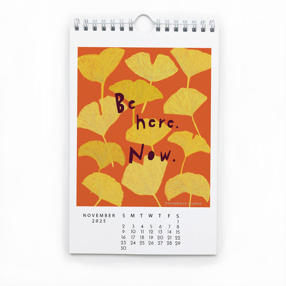 November 2025 calendar page with inspirational artwork featuring yellow ginkgo leaves on orange background and hand-lettered text "Be here. Now." Part of 2025 Follow Your Joy Monthly Wall Calendar.