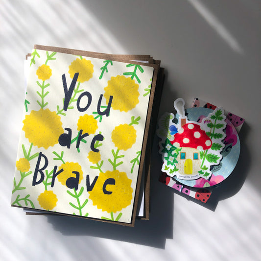 Colorful greeting card with yellow flowers and "You are Brave" text, alongside a cute mushroom sticker and other stationery items, showcasing the Seconds Mystery Pack contents