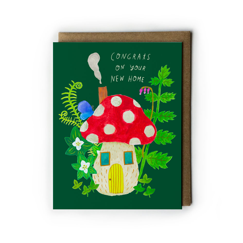 Whimsical greeting card featuring a colorful mushroom house with red cap and white spots, surrounded by green foliage, with "Congrats on your new home" text on dark green background, paired with kraft paper envelope