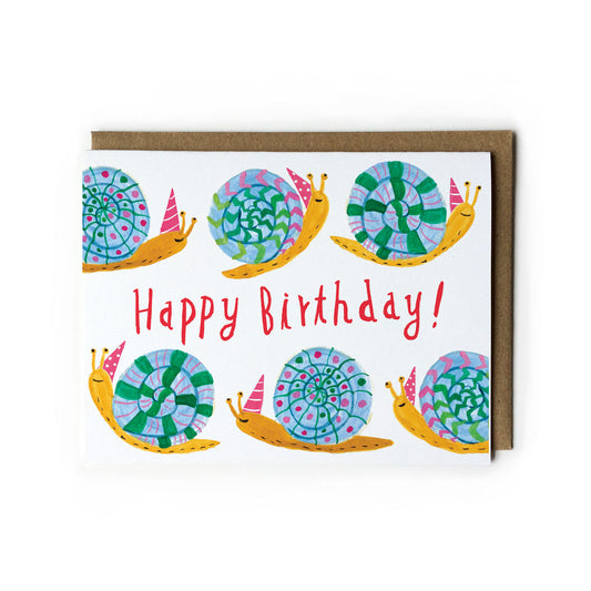 Colorful birthday card featuring six cheerful snails with vibrant patterned shells in blue, green, and purple. Text reads "Happy Birthday!" in red. Card has a white background and brown kraft envelope.