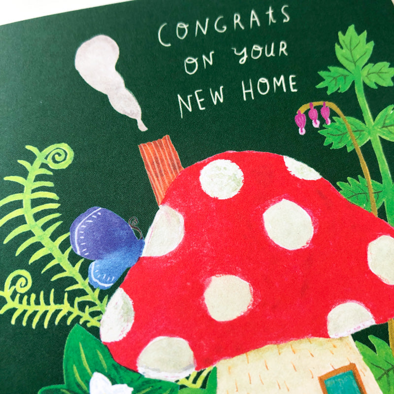 Whimsical greeting card with red and white spotted mushroom house, ferns, and butterfly on dark green background. Text reads "Congrats on your new home" in white handwritten style.