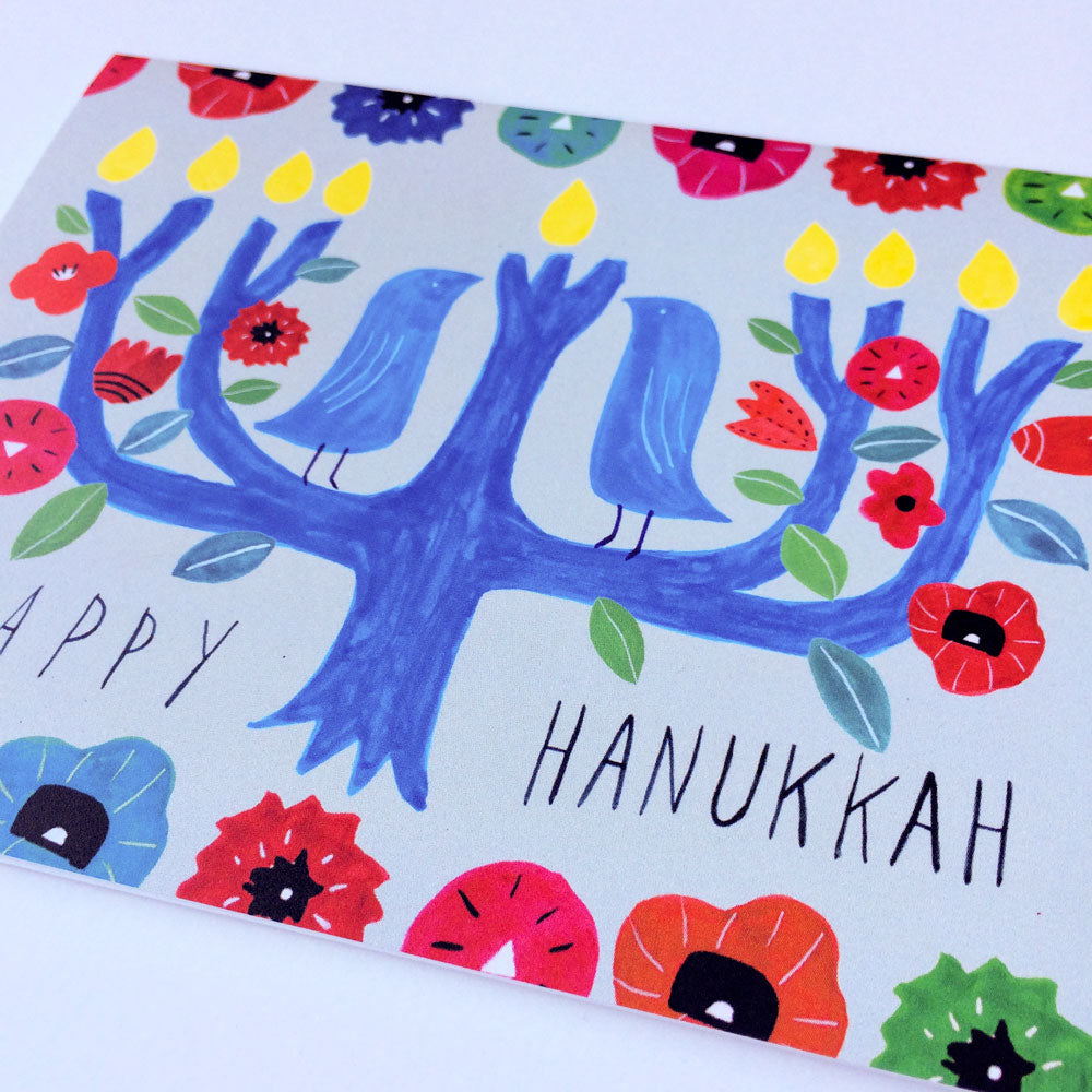 Whimsical Hanukkah card featuring a blue menorah tree with birds, colorful flowers, and handwritten "Happy Hanukkah" message. Vibrant watercolor design with festive floral elements surrounding the central tree shape, creating a cheerful and artistic holiday greeting.