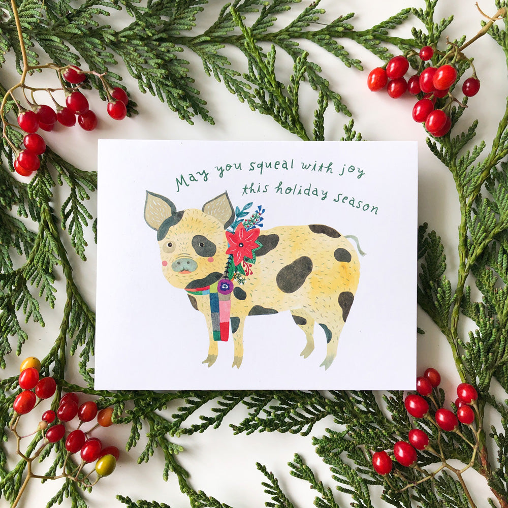 Holiday greeting card featuring watercolor illustration of spotted pig with festive flower, surrounded by evergreen branches and red berries, with handwritten message "May you squeal with joy this holiday season