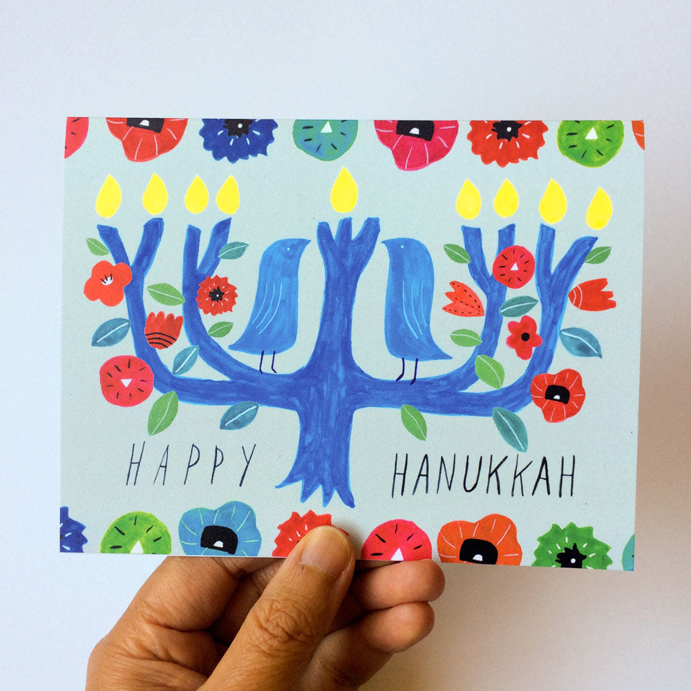 Colorful whimsical Hanukkah card featuring a blue menorah tree with yellow candles, surrounded by vibrant flowers and two stylized birds. Hand-drawn "Happy Hanukkah" message appears below the design. Card is held by a hand against a white background.
