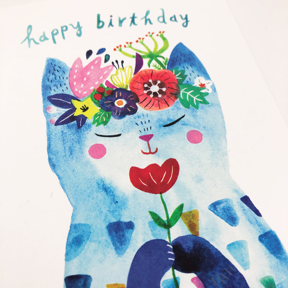 Whimsical watercolor illustration of a blue cat holding a red flower, wearing a colorful floral crown, with 'happy birthday' text above, on a white greeting card