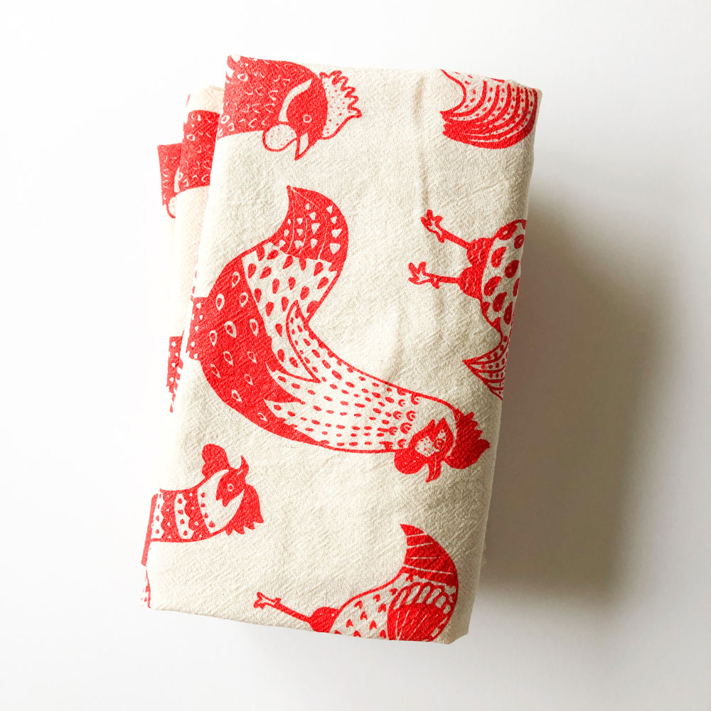 Folded cream-colored flour sack tea towel with red rooster pattern, featuring whimsical rooster designs in various poses and sizes, perfect for kitchen decor or as a unique gift.