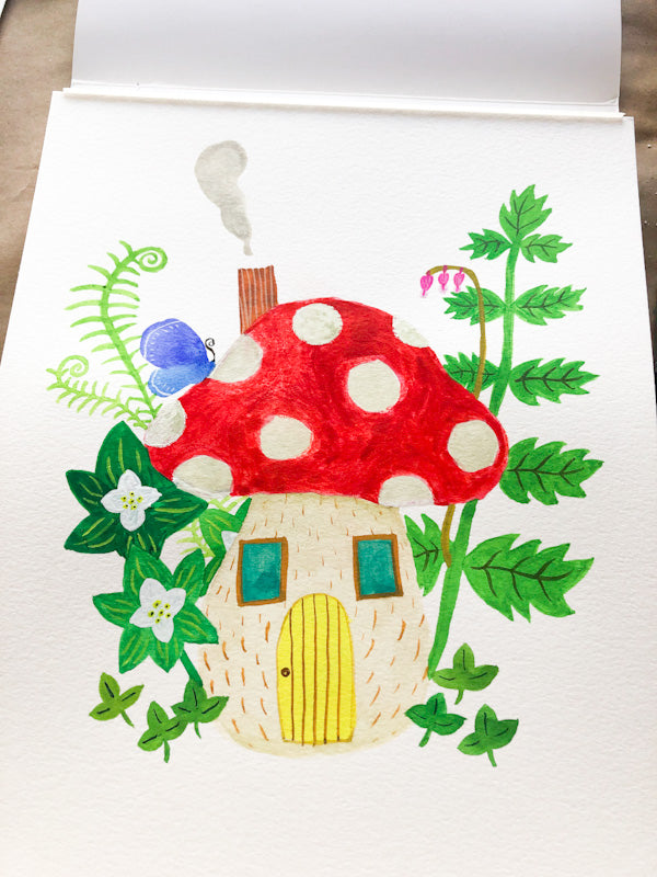 Whimsical watercolor illustration of a red and white polka dot mushroom house with yellow door, surrounded by green ferns, flowers, and leaves, on a white greeting card